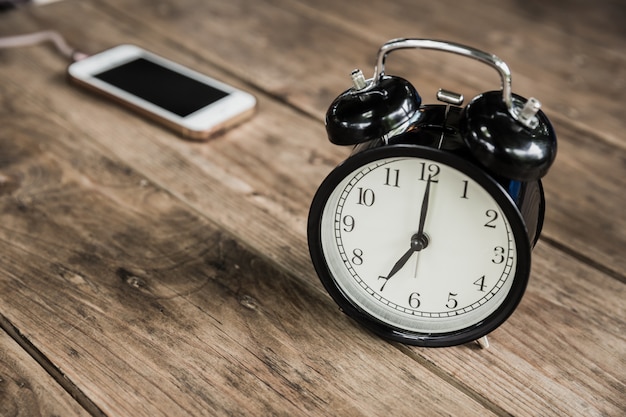 Las horas de reloj a las 7 de la mañana en la mesa de madera con un teléfono inteligente que se carga en el fondo borroso del café.