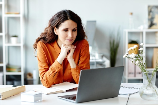 Horizontal de mujer gerente sentada en el escritorio en la oficina moderna viendo algo en internet en la computadora portátil