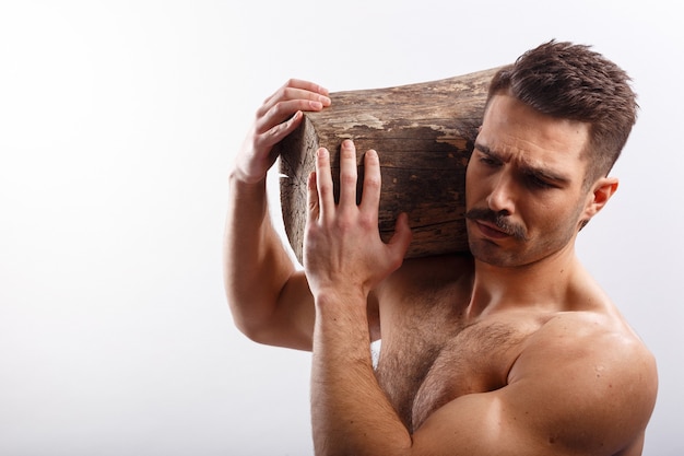 Horizontal retrato de un atleta, sexy, abs brutal barbudo con corte de pelo, con el torso desnudo, sosteniendo un tronco de árbol sobre sus hombros, sobre fondo blanco.