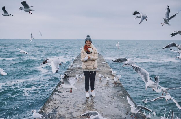 Horizontale Ansicht der jungen glücklichen Frau füttert Möwen auf dem Meer Hübsche Frau trägt Mantelschal und beobachtet fliegende Möwen am Meer am Himmel Menschen reisen Umwelt Naturkonzept