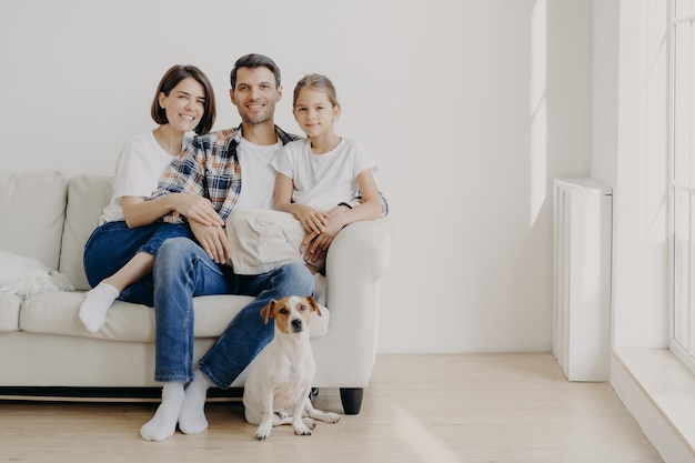 Horizontale Ansicht von drei freundlichen Familienmitgliedern und einem kleinen Rassehund, die vor der Kamera posieren, um unvergessliche Fotos zu machen Dauther verbringt Freizeit mit Mutter und Vater und hat einen guten Tag zu Hause