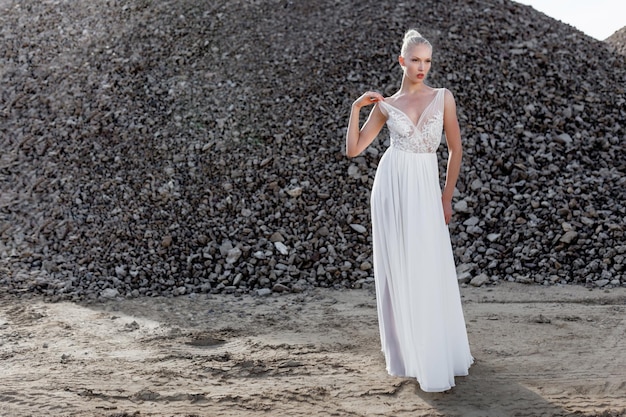 Horizontale Ansicht. Vorderes Bild der blonden jungen Frau in einem weißen Hochzeitskleid, die ihre Schulter berührt und tagsüber hinter Steinhaufen posiert.