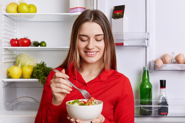 Horizontale Aufnahme der fröhlichen Frau hält Schüssel mit frischem Gemüsesalat, steht in der Nähe von Friedge voller Produkte, hält Diät. Vegetarierin isst nur Rohprodukte. Konzept für gesunde Ernährung