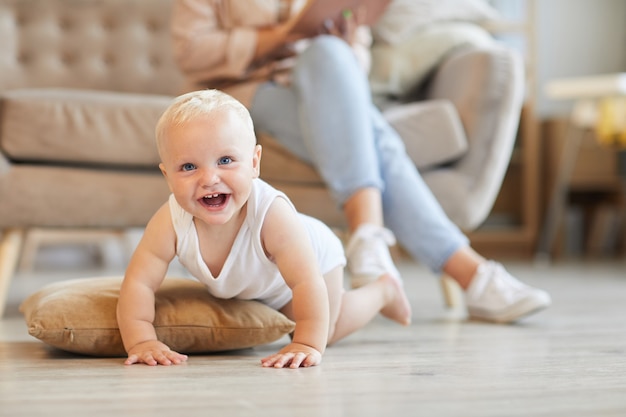 Horizontale Aufnahme des verspielten Babys, das Spaß auf Boden im Wohnzimmer hat, während seine Mutter auf Sofa sitzt und Buch liest