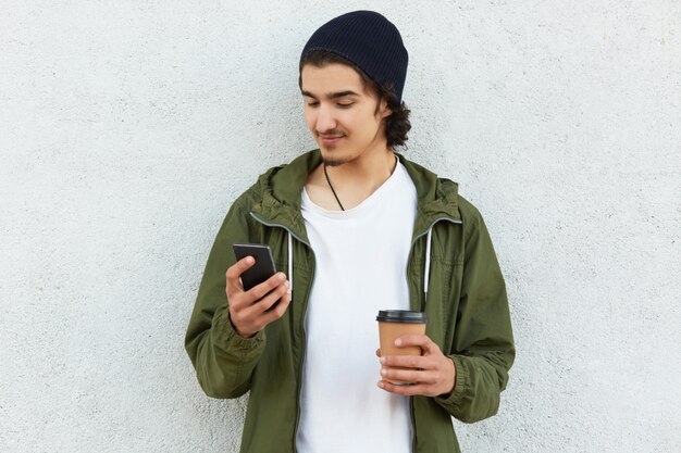 Horizontale Aufnahme des zufriedenen konzentrierten Mannes trägt stilvolle Kopfbedeckungen, trägt Kaffee zum Mitnehmen, surft in sozialen Netzwerken auf dem Handy