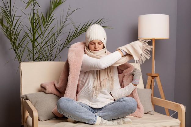 Horizontale aufnahme einer attraktiven gefrorenen frau mit mütze, handschuhen, schal und mantel, die auf dem sofa im wohnzimmer sitzt und keine zentralheizung in ihrer wohnung hat, die versucht, sich aufzuwärmen