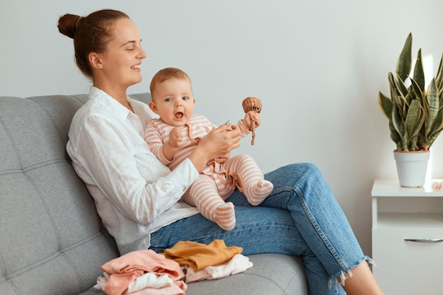 Horizontale Aufnahme einer attraktiven, schönen Mutter, die mit ihrer kleinen Tochter auf Husten sitzt und dem Kind Holzspielzeug zeigt, eine Frau mit weißem Hemd und Jeans.