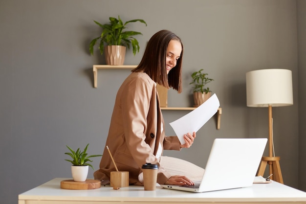 Horizontale Aufnahme einer attraktiven, zufriedenen kaukasischen Frau mit braunen Haaren, die eine beige Jacke trägt, die im Büro arbeitet, Dokumente hält und auf einem Notizbuch tippt