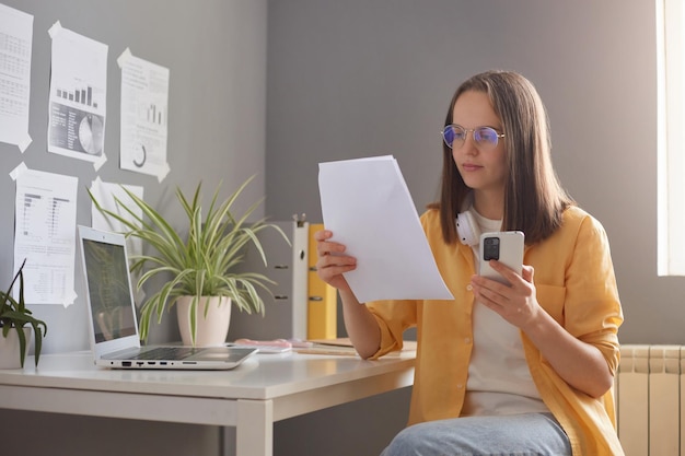 Horizontale Aufnahme einer braunhaarigen Frau mit Fokus auf eine Frau mit gelbem Hemd, die im Büro an ihrem Arbeitsplatz sitzt und Dokumente und ein Handy in den Händen hält, die etwas auf Papier lesen