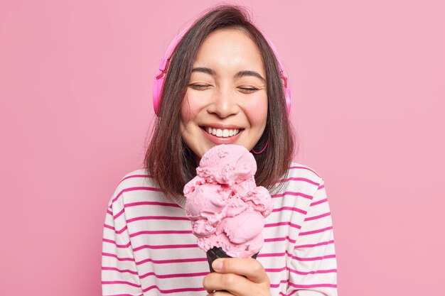 Horizontale Aufnahme einer fröhlichen, schönen asiatischen Jahrtausendfrau schließt die Augen vor Vergnügen, während sie leckeres süßes Eis isst, wie Sommerdesser Musik hört, drahtlose Kopfhörer verwendet, genießt das Wochenende