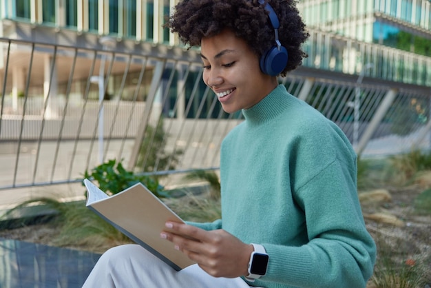 Horizontale Aufnahme einer glücklichen Studentin mit glücklichem Lächeln lernt Informationen aus persönlichen Notizen hält Notizbuch verwendet Stereokopfhörer hört Audiospur in Freizeitkleidung Posen im Freien