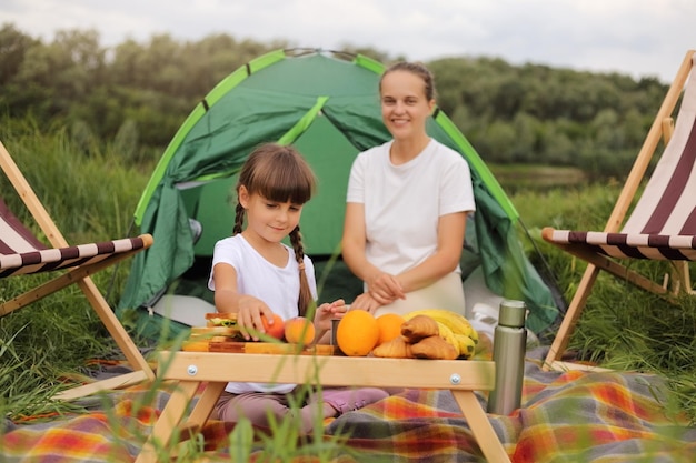 Horizontale Aufnahme einer gut aussehenden Frau mit ihrer Tochter in der Nähe des Zeltes am Holztisch voller Essen, die das Camping in der Nähe des Flusses genießt und Obst und Sandwiches isst