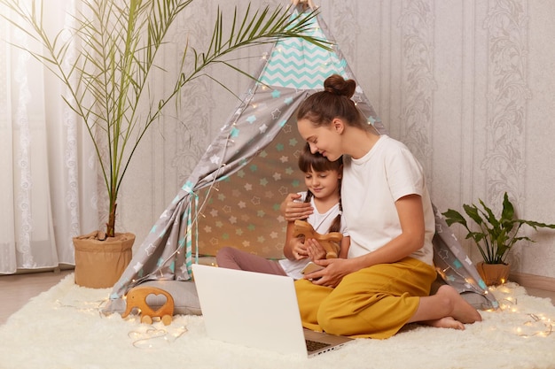 Horizontale Aufnahme einer hübschen Frau und ihrer Tochter in weißen T-Shirts, die im Wigwam vor dem Laptop sitzt und gemeinsam Cartoons anschaut Mama umarmt ihr süßes Kind