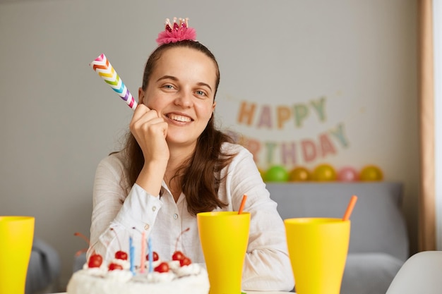 Horizontale Aufnahme einer jungen erwachsenen entzückenden Frau mit weißem Hemd, die Geburtstag feiert, allein am Tisch mit Kuchen sitzt und ein Partyhorn hält, das lächelnd in die Kamera schaut