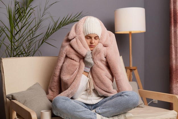 Horizontale Aufnahme einer kalten Frau, die zu Hause auf dem Sofa sitzt, mit gekreuzten Beinen, Hut und mit Fäustlingen umwickelten Wintermantel, die versucht, sich in einer gefrorenen Wohnung ohne Zentralheizung aufzuwärmen