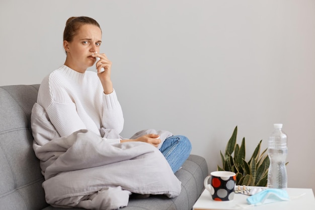 Horizontale Aufnahme einer kranken Frau, die in eine Decke gehüllt auf dem Sofa sitzt, sich erkältet, sich schlecht fühlt, unter Kopfschmerzen und hohen Temperaturen leidet, Grippesymptome.
