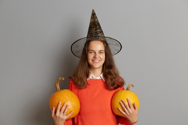 Horizontale Aufnahme einer lächelnden attraktiven Frau mit Pullover und Hexenhut, die mit orangefarbenem Kürbis in den Händen isoliert vor grauem Hintergrund steht und in die Kamera schaut, die in guter festlicher Halloween-Stimmung ist