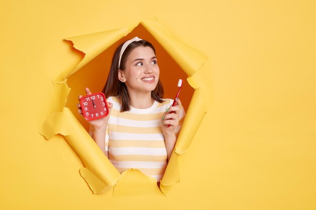 Horizontale Aufnahme einer lächelnden Frau mit gestreiftem Hemd und Haarband, die in gelbem Papierloch posiert und Wecker und Zahnbürste hält, die auf den Kopierraum für Werbung oder Promotion wegschaut