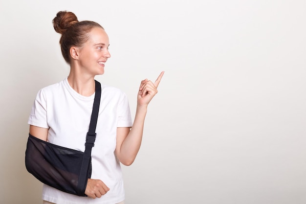 Horizontale Aufnahme einer lächelnden, positiven Frau mit Brötchenfrisur, die ein lässiges T-Shirt trägt und isoliert vor weißem Hintergrund posiert. Frau mit gebrochenem Arm in Schiene, die auf den Werbebereich zeigt