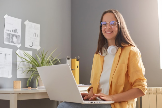 Horizontale Aufnahme einer lächelnden schönen Frau mit braunen Haaren, die ein gelbes Hemd trägt und im Büro posiert, das am Laptop arbeitet und mit einem breiten Lächeln in die Kamera blickt