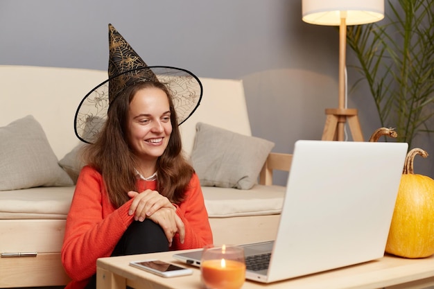 Horizontale Aufnahme einer lächelnden, zufriedenen kaukasischen Frau mit Hexenhut, die einen Videoanruf auf dem Laptop hat oder einen Livestream sendet, der mit positivem Ausdruck auf das Display blickt, um die Feiertage zu Hause zu feiern