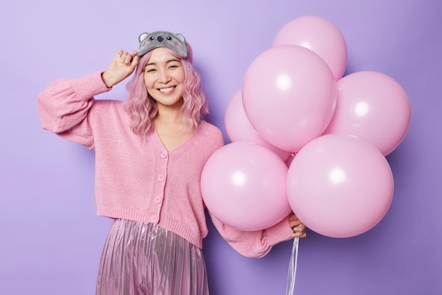 Horizontale Aufnahme einer positiven Frau, die breit lächelt, hat lang gefärbte rosa Haare, die in festliche Kleidung gekleidet sind, hält einen Haufen Heliumballons und verbringt gerne Freizeit auf einer Party, hat einen besonderen Anlass im Leben