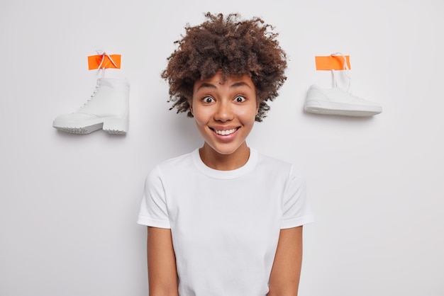 Horizontale Aufnahme einer positiven jungen Frau mit lockigem dunklem Haar lächelt glücklich in lässigen Basic-T-Shirt-Posen vor weißen Hintergrundschuhen, die an der Wand verputzt sind, wählt zwischen Stiefel und Sneaker