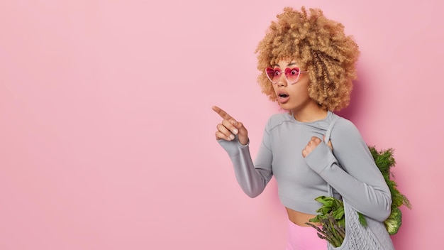 Horizontale Aufnahme einer schockierten, lockigen jungen Frau, die eine Tasche mit frischem Bio-Gemüse trägt, zeigt mit dem Zeigefinger auf den Kopienraum, der über rosafarbenem Hintergrund isoliert ist, genießt es, natürliche Lebensmittel zu essen