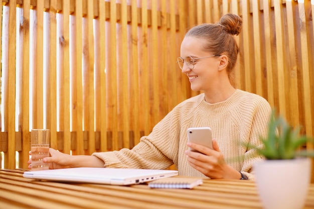 Horizontale Aufnahme einer schönen optimistischen Frau mit Brötchenfrisur, die einen beigen Pullover trägt und ein Handy in den Händen hält, hält ein Glas Wasser und fühlt sich durstig