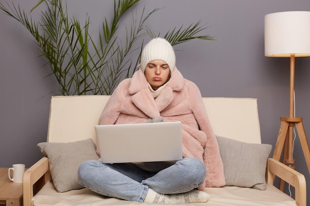 Horizontale Aufnahme einer traurigen kaukasischen Frau, die in einem kalten Raum posiert und auf dem Sofa sitzt und warme Mützen und Mützen trägt, die einen Laptop auf Knien halten und mit schmollenden Lippen auf das Display schauen