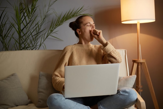 Horizontale Aufnahme einer verschlafenen Frau in Freizeitkleidung, die auf dem Sofa sitzt und an einem PC arbeitet, die müde ist und schlaflos gähnt und ihren Mund bedeckt