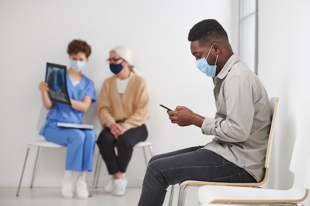 Horizontale Aufnahme eines afroamerikanischen Mannes mit Maske im Gesicht, der auf einem Stuhl im Krankenhauskorridor sitzt w