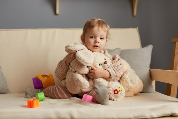 Horizontale Aufnahme eines charmanten blonden Babys, das auf Husten sitzt, mit ihren weichen Spielsachen, die ihr Kaninchen und ihren Teddybären umarmen und in die Kamera schauen, während sie ihre Freizeit zu Hause verbringen
