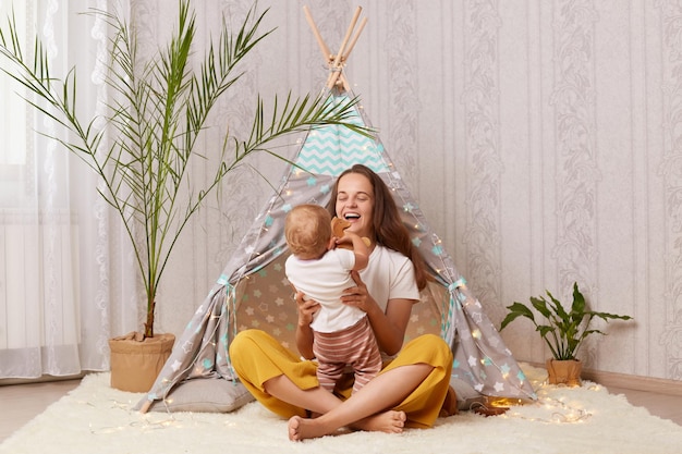 Horizontale Aufnahme eines glücklichen Babymädchens und ihrer Mutter, die zu Hause in Wigwam spielen, sich umarmen und lächeln und glücklich und zufrieden sind, wenn sie das Wochenende zusammen verbringen