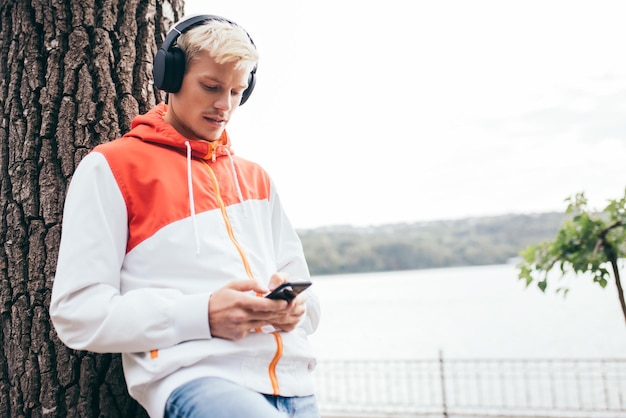 Horizontale Aufnahme eines jungen blonden Mannes, der das Wetter im Stadtpark genießt und auf seinen Freund wartet, während er seinen Lieblingstrack mit dem Handy im Freien im Park hört Lifestyle-Konzept