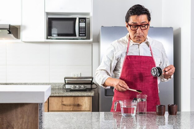 Horizontale Aufnahme eines lateinamerikanischen Mannes mit roter Schürze, der gemahlenen Kaffee in eine französische Presse neben zwei Espressotassen in einer Küche stellt