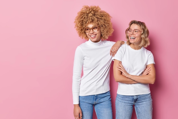 Foto horizontale aufnahme von glücklichen jungen frauen, die gerne lächeln, tragen legere kleidung haben unbeschwerte ausdrücke, die vor rosa hintergrund stehen, leerer kopierraum für ihre werbeinhalte freundschaftskonzept