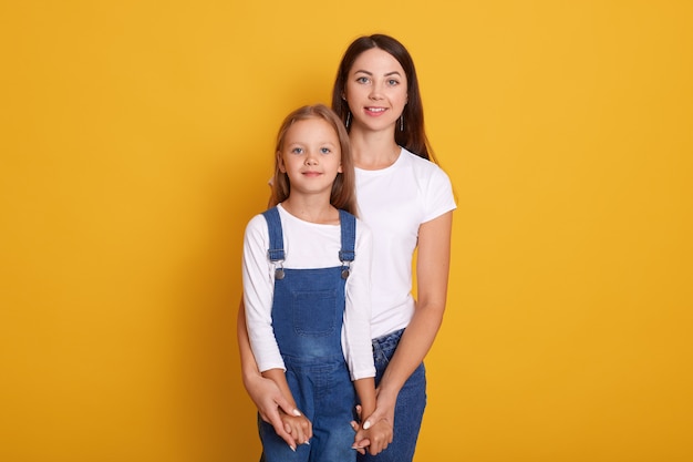 Horizontale Aufnahme von Mutter mit Tochter, glücklich zusammen verbringen, weibliche Kleider weißes T-Shirt, niedliches kleines Mädchen, das Overalls trägt