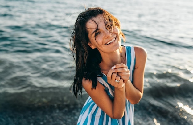 Horizontale Bild von glücklich Brünette schöne Frau mit Wind weht Haar posiert auf dem Meer auf Sonnenuntergang Hintergrund Kaukasische schöne Mädchen lächelnd glücklich am Strand Urlaub genießen warmen Sonnenschein