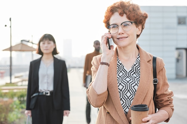 Horizontale mittlere Porträtaufnahme einer schönen reifen Frau mit lockigen roten Haaren, die nach Beendigung des Arbeitstages mit jemandem auf dem Heimweg telefoniert