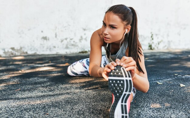 Foto horizontales bild der jungen fitnessfrau, die sich ausdehnt und die musik auf kopfhörern hört, bevor sie im freien gegen die betonwand athletische weibliche streckung nach dem training außerhalb des sport- und personenkonzepts