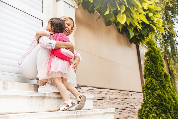 Horizontales Bild einer glücklichen Mutter mit Treffen mit ihrer Schülertochter, die vom Kindergarten nach Hause zurückkehrt Niedliches kleines Mädchen mit Mutter umarmt sich nach dem ersten Vorschultag glücklich Familie