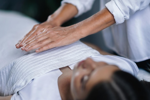 Horizontales Bild einer weiblichen Reiki-Therapeutin, die die Hände über die Patientenbrust hält, die das Herzchakra heilt, ein friedliches, schönes Teenagermädchen, das mit geschlossenen Augen liegt