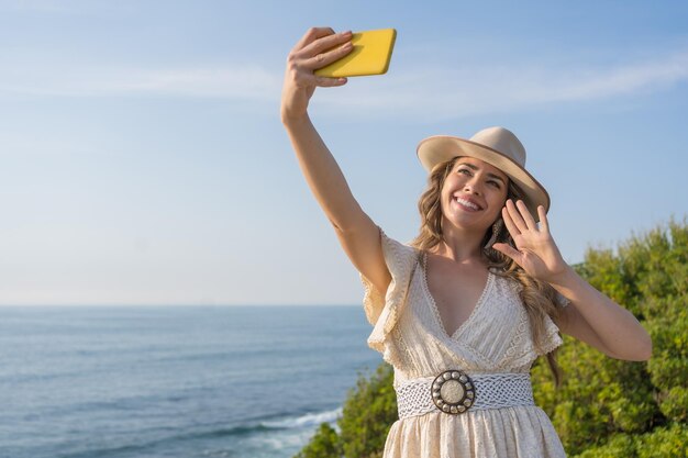 Foto horizontales foto einer blonden schönheitsfrau in sommerkleidung, die beim selfie am meer winkt
