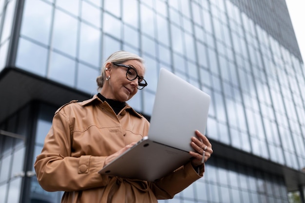 Horizontales Foto einer erfolgreichen, reifen, erwachsenen Chefin mit Laptop auf Gebäudehintergrund