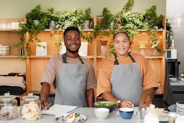 Horizontales mittleres Porträt junger afroamerikanischer Männer und Frauen, die in der Café-Küche arbeiten und ein neues Gericht kreieren, das Rezept mit Blick auf die Kamera aufschreibt