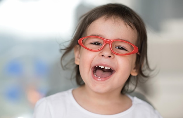 Horizontales Nahaufnahmeporträt von intelligenten, glücklichen, kaukasischen, süßen, kleinen Studentenmädchen, die lachen Neugieriges, glückliches, schönes Kleinkindmädchen mit roter Brille Bildungskonzept