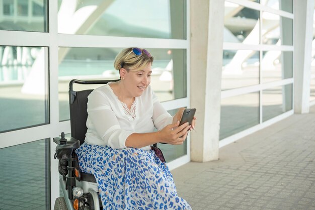 Horizontales Porträt einer lächelnden kaukasischen Frau im Rollstuhl, die auf ein Handy blickt