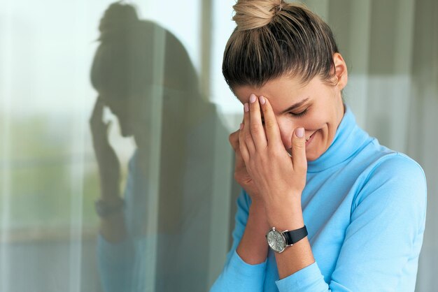 Horizontales Porträt einer schönen jungen Frau, die neben dem Fenster im Büro lächelt und posiert und einen blauen Rollkragenpullover trägt, wobei die Finger ihre Stirn berühren Menschen- und Geschäftskonzept