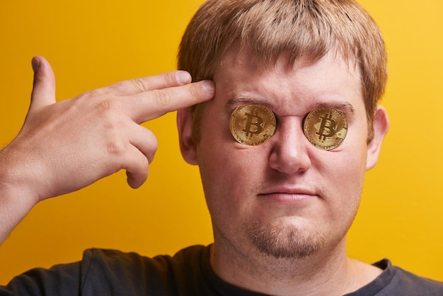 Horizontales Porträt eines dicken Kerls mit Bitcoins in den Augen auf gelbem Hintergrund. Digitales virtuelles Währungskonzept, betrogener Investor, Kryptowährung fallen auf den Internetmarkt